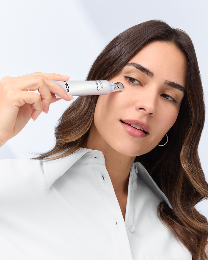 Ein weibliches Model trägt das ALCINA Augenfluid Koffein Booster auf die Haut unter den Augen auf. Die Szene zeigt eine Nahaufnahme ihres Gesichts mit sichtbarer Produktflasche in der Hand. Das Fluid steht für eine belebende Augenpflege gegen Müdigkeitsanzeichen, verpackt in einem schlichten, modernen Design