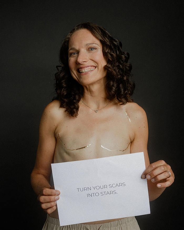 Portrait einer Frau ohne Brüste, die Narben sind gold bemalt. Sie hält eine Nachricht ind er Hand für das Brustkrebs "Let your scars shine" Fotoprojekt von Paul Mitchell