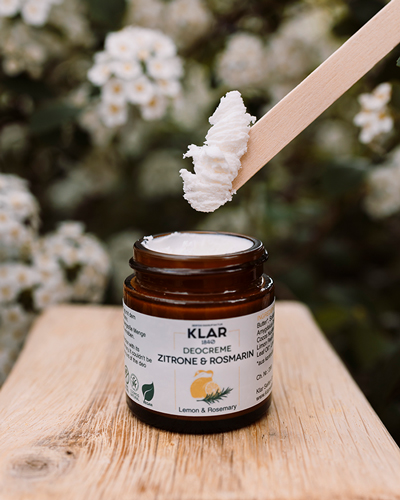 Nachhaltige Deocreme im Glas von Klar Seifen, Sorte Zitrone & Rosmarin, cremige Textur auf Holzuntergrund, im Hintergrund weiße Blüten – natürliche und umweltfreundliche Körperpflege.